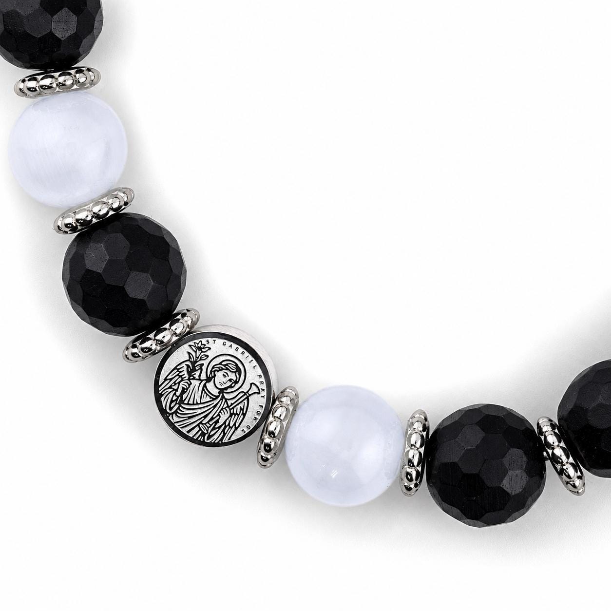 Close-up view of a beaded bracelet showing faceted black onyx beads, smooth white selenite beads, and stainless steel spacers arranged in a curved section. At the center is a round stainless steel charm engraved with Archangel Gabriel and his sigil. The detailed etching, bead textures, and polished metal accents are clearly visible against a bright white background.